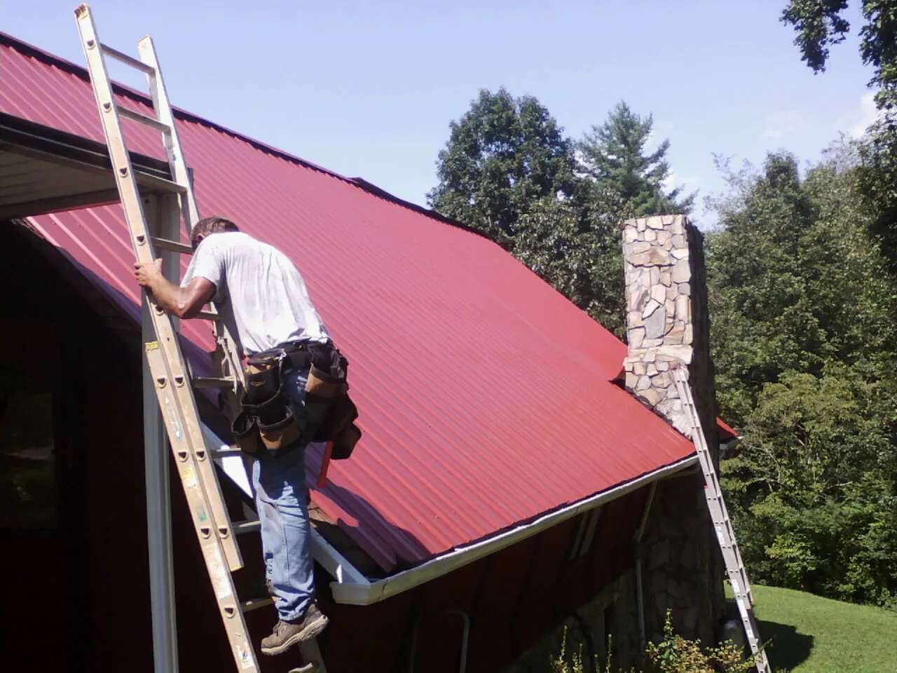 Roofer on a ladder beside a red metal roof and stone chimney — Soffit & Fascia Repair by Roofing Brothers in Hemlock